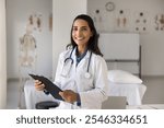 Beautiful Latina female cardiologist holding clipboard posing for camera standing alone in modern examination room. Portrait of professional medical worker, medicine, mission, achievements and career