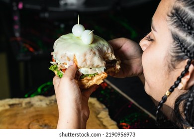 Beautiful Latina Girl With Brown Skin And Braids In Her Hair, Eating A Delicious Colombian Hamburger Covered With Melted Cheese In The Streets Of Colombia. Hamburger Bite.