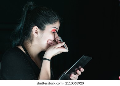 beautiful latina woman with green hair, making up her eyes with a red eye shadow. girl holding her brush while putting it on her right eye. black background. beauty concept.