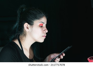 beautiful latina woman with green hair, making up her eyes with a red eye shadow. girl holding the mirror of her eye shadow palette while observing if she looks good. black background. beauty concept.