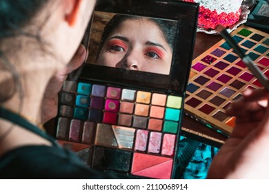 Beautiful Latina woman looking in the mirror of her eye shadow palette, with a palette of colored shadows in the background on top of her dressing table, girl doing her eye makeup with red color.
