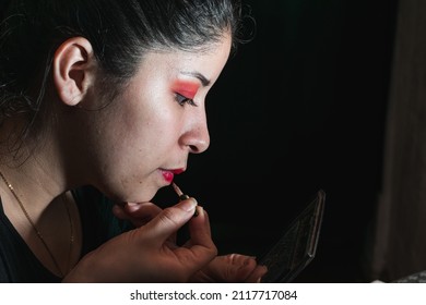 beautiful latina woman using a brush to paint her lips red, opening her mouth a little to apply the lipstick carefully. her eyes are painted with a red shadow. black background.