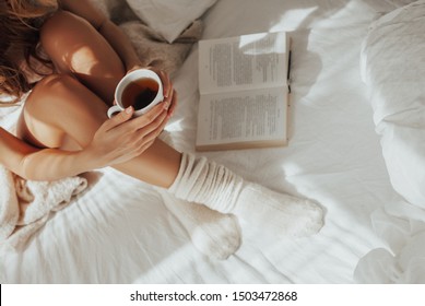 Beautiful Legs on Bed and White Linens in Silk, Woman with a Cup of Refreshing Tea, Warm Knitted Socks, she is basked in the Bed and Reading a Book, the sun's rays shine into  the        - Powered by Shutterstock