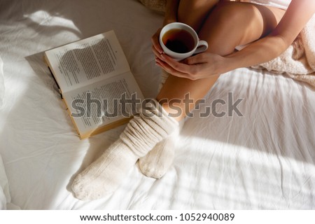 Beautiful Legs on Bed and White Linens in Silk, Woman with a Cup of Refreshing Tea, Warm Knitted Socks, she is basked in the Bed and Reading a Book, the sun's rays shine into the Bedroom            