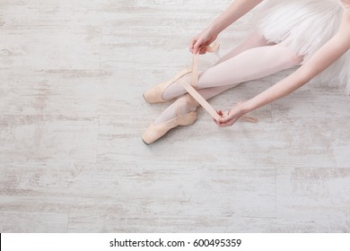 Beautiful legs of young ballerina who puts on pointe shoes at white wooden floor background, top view from above with copy space. Ballet practice. Beautiful slim graceful feet of ballet dancer.