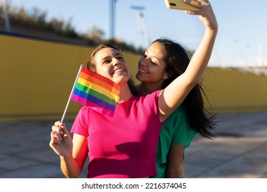 A Beautiful Lesbian Young Couple Embraces And Holds A Rainbow Flag. Girls Taking Selfie Photo