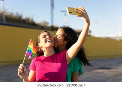  A Beautiful Lesbian Young Couple Embraces And Holds A Rainbow Flag. Girls Taking Selfie Photo