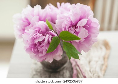 Beautiful lifestyle photo of freshly cut pink peonies in a vintage copper vase in front of a sunlit window old wooden board and linen runner in the background - Powered by Shutterstock