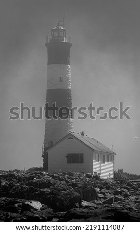 Beautiful lighthouse on the top of a rocky mountain with sea in foggy morning in black and white photo. Lighthouse on black rocks among the fog in the sea. Dramatic seascape, nobody, travel photo