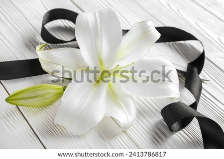Beautiful lily flowers and black funeral ribbon on white wooden background
