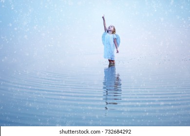 Beautiful little angel girl pointing a finger at the sky and standing knee-deep in water reflected in the water surface. Snowflakes background.