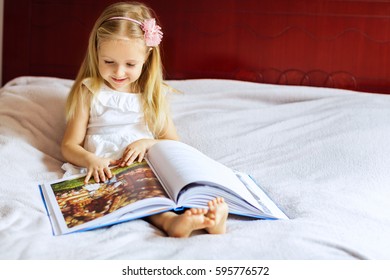 Beautiful Little Blond Girl Sitting At Home On The Bed And Reading A Children's Book