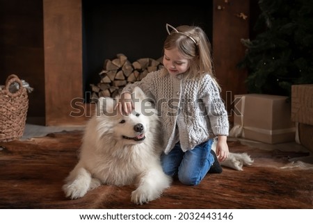 Beautiful little blonde baby girl, has happy fun face, pretty eyes, embrace samoyed husky dog puppy. Portrait holiday home. Winter family animal. Kids fashion style. 