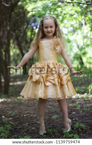 A beautiful little blonde girl in a golden dress with the crown on her head is alone in the forest