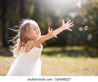 Beautiful little blonde girl, has happy fun cheerful smiling face, white dress, soap bubble blower. Portrait nature. 