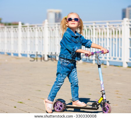 Beautiful little blonde girl, has happy fun cheerful smiling face, blue jeans and sunglasses. Motion on great kick scooter in urban city. Portrait nature. 