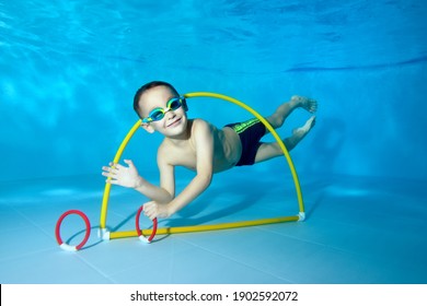 A Beautiful Little Boy Dives Underwater In A Children's Pool. He Swims Through The Hoop, Pulls Out Toys. Active Happy Child. Healthy Lifestyle. Swimming Lessons Under The Water. A Family Sport