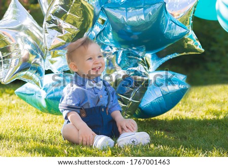 A beautiful little boy kid gentleman in a retro suit with suspenders and a bow tie is sitting on a green lawn. Children's party with balloons happy birthday, 1 year.