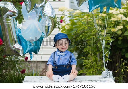 A beautiful little boy kid gentleman in a retro suit with suspenders and a bow tie is sitting on a white tablen. Children's party with balloons happy birthday, 1 year.
