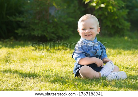 A beautiful little boy kid gentleman in a retro suit with suspenders and a bow tie is sitting on a green lawn. Children's party happy birthday, 1 year.