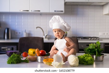 Beautiful little boy,has happy fun smiling face, white chef hat. Cooks in kitchen appetizing tasty vegetables. Child portrait. Detox concept. Cuisine food view. Home interior.