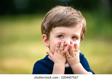 A Beautiful Little Caucasian Boy, Looking Scared Or Guilty, Covers His Face With His Hands, Hiding, In A Park, As If To Apologize After Having Made A Little Mistake. Concept Of Emotions In Children.