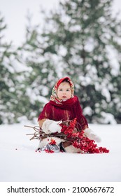 Beautiful little child girl plays by viburnum bunches in snowy forest. She dressed in the old Russian style in red headscarf.