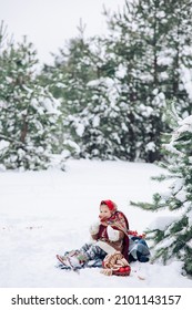 Beautiful little child girl plays in snowy forest near basket with apples and bagels. She dressed in the old Russian style in red headscarf.