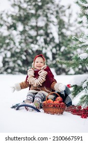 Beautiful little child girl sits with bagels bunch on her neck near basket of apples during the walk in snowy forest. She dressed in the old Russian style in red headscarf.