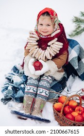 Beautiful little child girl sits with bagels bunch on her neck near basket of apples during the walk in snowy forest. She dressed in the old Russian style in red headscarf.