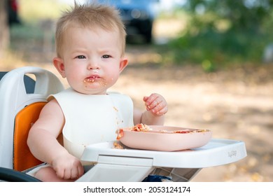 A beautiful little child is having breakfast. A dirty child sits at a table in nature. The baby sits at the children's table, takes food from the plate with her hands and eats. Breakfast