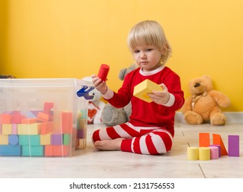 A beautiful little child in red pajamas is sitting on floor in playroom and playing with colorful cubes of figures folded in a transparent box. Selective focus