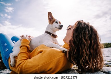 A Beautiful Little Dog Sitting On Its Owner Looking At Her Face. The Woman Is Lying Down In The Park In A Sunny Day In Madrid. Family Dog Outdoor Lifestyle