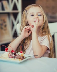 Beautiful Little Girl Blonde Woman Smiling In Cafe Eats A Dessert Ice Cream With Fruits ,close-up Holds The Index Finger In The Mouth,the Concept Of Holidays And Weekends.A Happy Childhood