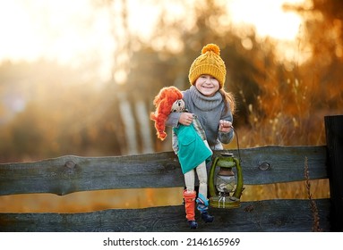 Beautiful little girl in a cap plays with pippi longstocking. Girl on the fence with a doll and a flashlight laughs and smiles at the camera.