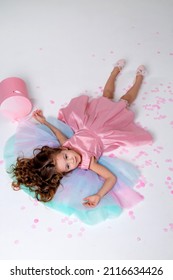 Beautiful Little Girl In A Chic Pink Dress Lies On The Floor Strewn With Confetti. Top View. Fashion And Style. Child Celebrates His Birthday. Photo In The Studio. Space For Text. High Quality Photo