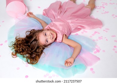 Beautiful Little Girl In A Chic Pink Dress Lies On The Floor Strewn With Confetti. Top View. Fashion And Style. Child Celebrates His Birthday. Photo In The Studio. Space For Text. High Quality Photo