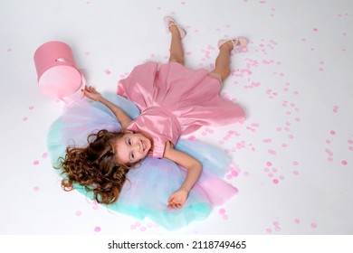 Beautiful Little Girl In A Chic Pink Dress Lies On The Floor Strewn With Confetti. Top View. Fashion And Style. Child Celebrates His Birthday. Photo In The Studio. Space For Text. High Quality Photo