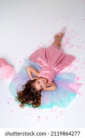 Beautiful Little Girl In A Chic Pink Dress Lies On The Floor Strewn With Confetti. Top View. Fashion And Style. Child Celebrates His Birthday. Photo In The Studio. Space For Text. High Quality Photo