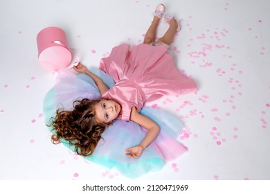 Beautiful Little Girl In A Chic Pink Dress Lies On The Floor Strewn With Confetti. Top View. Fashion And Style. Child Celebrates His Birthday. Photo In The Studio. Space For Text. High Quality Photo