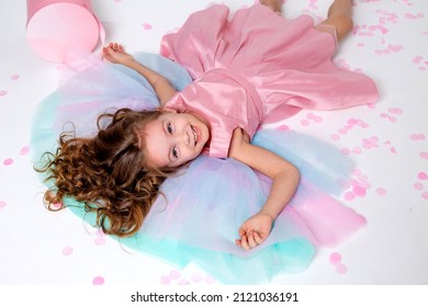 Beautiful Little Girl In A Chic Pink Dress Lies On The Floor Strewn With Confetti. Top View. Fashion And Style. Child Celebrates His Birthday. Photo In The Studio. Space For Text. High Quality Photo