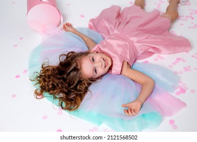 Beautiful Little Girl In A Chic Pink Dress Lies On The Floor Strewn With Confetti. Top View. Fashion And Style. Child Celebrates His Birthday. Photo In The Studio. Space For Text. High Quality Photo