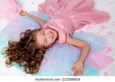 Beautiful Little Girl In A Chic Pink Dress Lies On The Floor Strewn With Confetti. Top View. Fashion And Style. Child Celebrates His Birthday. Photo In The Studio. Space For Text. High Quality Photo