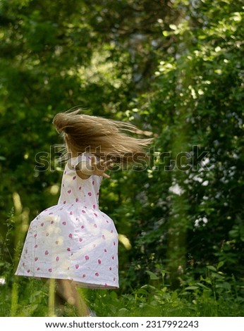 A beautiful little girl dances and spins happily and cheerfully in the park in the summer. Light long hair is developing. Against the background of green trees.