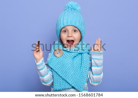 Beautiful little girl expressing joyous emotions, keeps ahnds up and mouth opened, child wearing casual striped shirt, scarf and cap with pom pom, holding candy in hand, isolated over blue background.