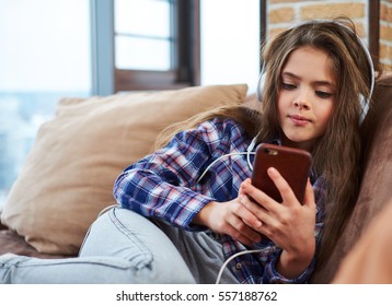 Beautiful little girl in headphones using smartphone on a couch