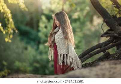 A beautiful little girl with long blond hair wrapped in a knitted blanket stands in the forest on a hill. A girl stands thoughtfully near a tree branch in the park. 