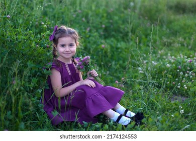 A beautiful little girl with long hair, in a purple dress, smiles and looks at the camera, holding a bouquet of clover in her hands