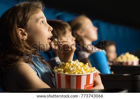 Beautiful little girl looking fascinated eating popcorn watching a movie at the local movie theatre snack bucket junk food tasty childhood entertaining entertained emotions kids concept