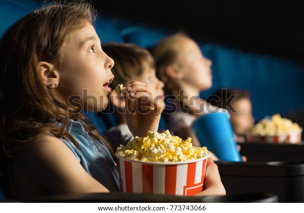 Beautiful\
little girl looking fascinated eating popcorn watching a movie at\
the local movie theatre snack bucket junk food tasty childhood\
entertaining entertained emotions kids\
concept